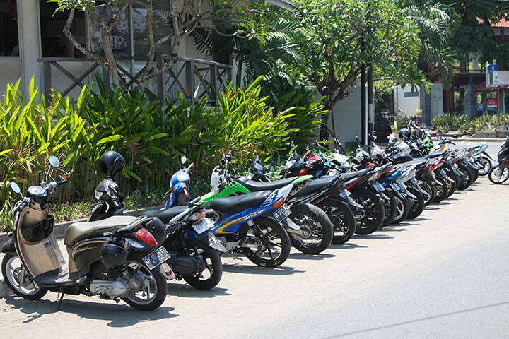  Many motorbikes are parked in a row on a beautiful, Sunny, green street in Seminyak. You can rent them to see the surrounding attractions.