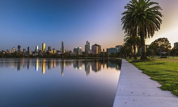 Melbourne city skyline illuminated at sunset, reflecting in the lake, with a peaceful footpath in the foreground.