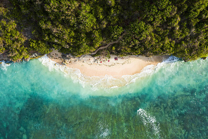 View of Green Bowls Beach, Bali, from above,  featuring a stunning aerial view of some tourists sunbathing on a beautiful beach bathed by a turquoise rough sea during sunset